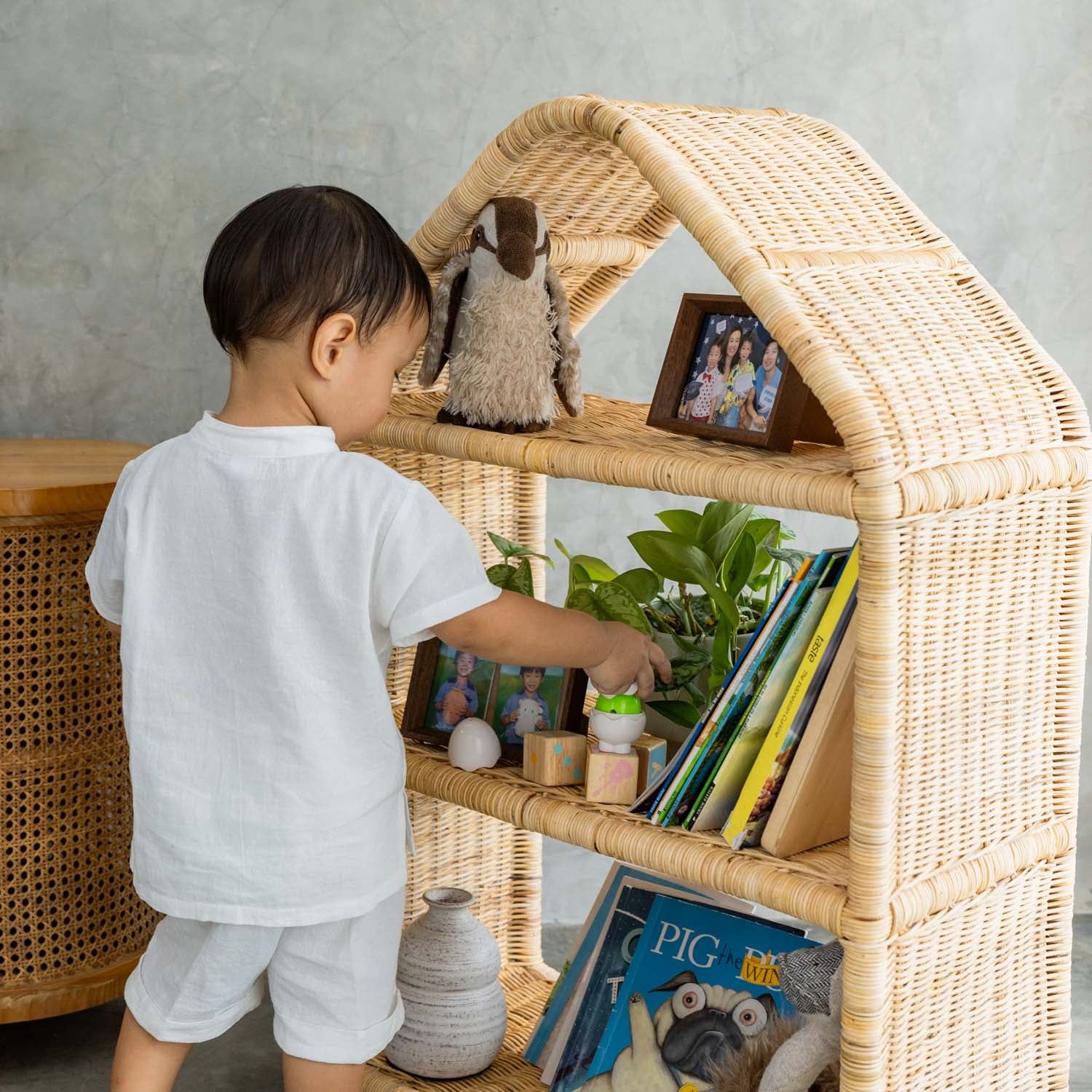 Child interacting with the handcrafted rattan wicker Alaya house shelf by MOMIJI, filled with children’s books and toys in a calm indoor setting.