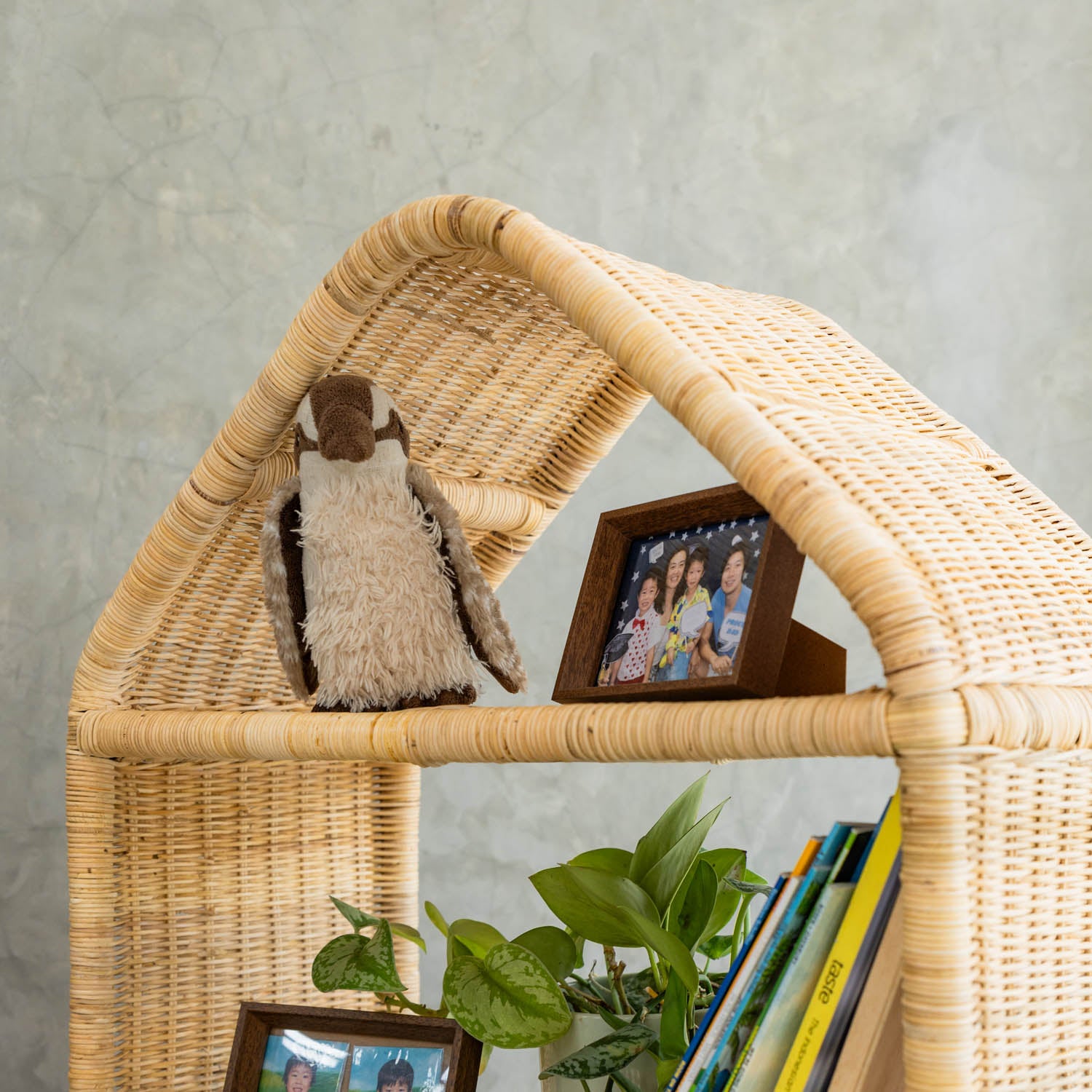 Detail of the top section of the handcrafted Alaya house shelf by MOMIJI made from rattan wicker, featuring a penguin doll and framed photos styled in beige neutral color.