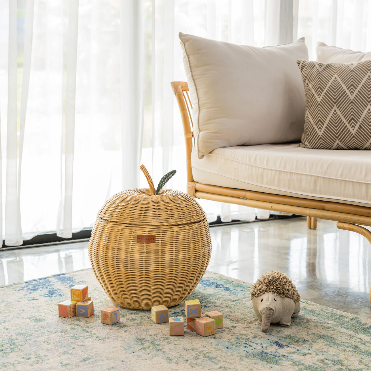 Handcrafted natural beige rattan wicker apple storage basket in medium size by MOMIJI, styled on a rug with children’s toys, featuring a green leaf and natural stem detail on the lid.
