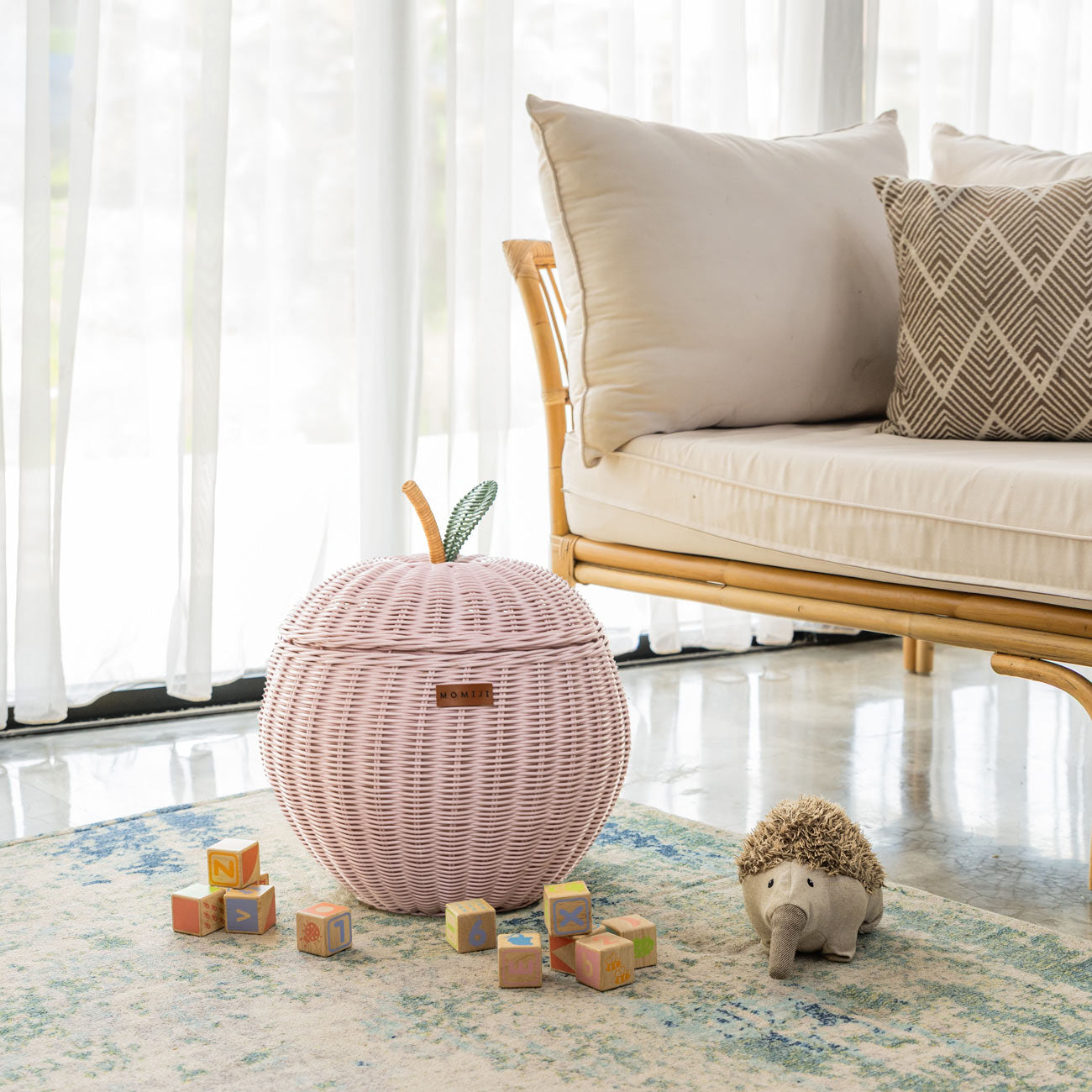 Handcrafted pink rattan wicker apple storage basket in medium size by MOMIJI, styled on a rug with children’s toys, featuring a green leaf and natural stem lid detail.