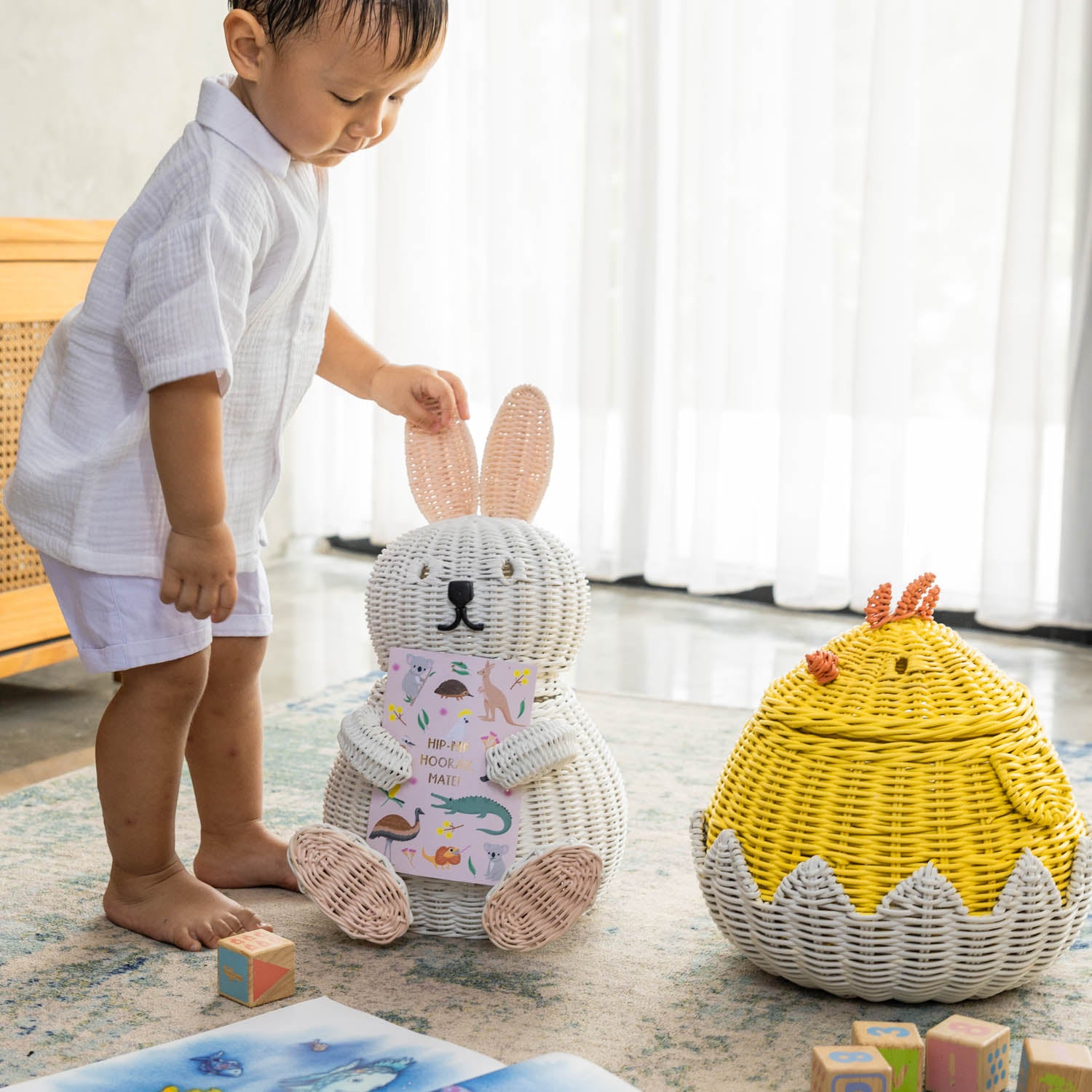 Child holding a handcrafted white rattan wicker bunny storage basket by MOMIJI in a room styled with a chick basket, toys, and children’s books.
