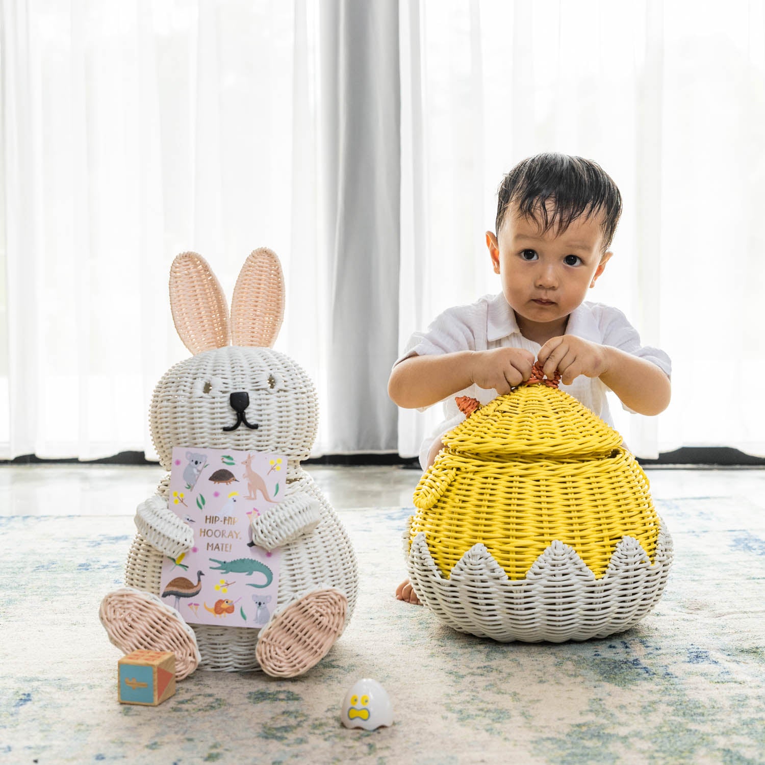 Handcrafted white rattan wicker bunny storage basket by MOMIJI styled holding children’s books on a rug, with a child nearby holding a chick basket.