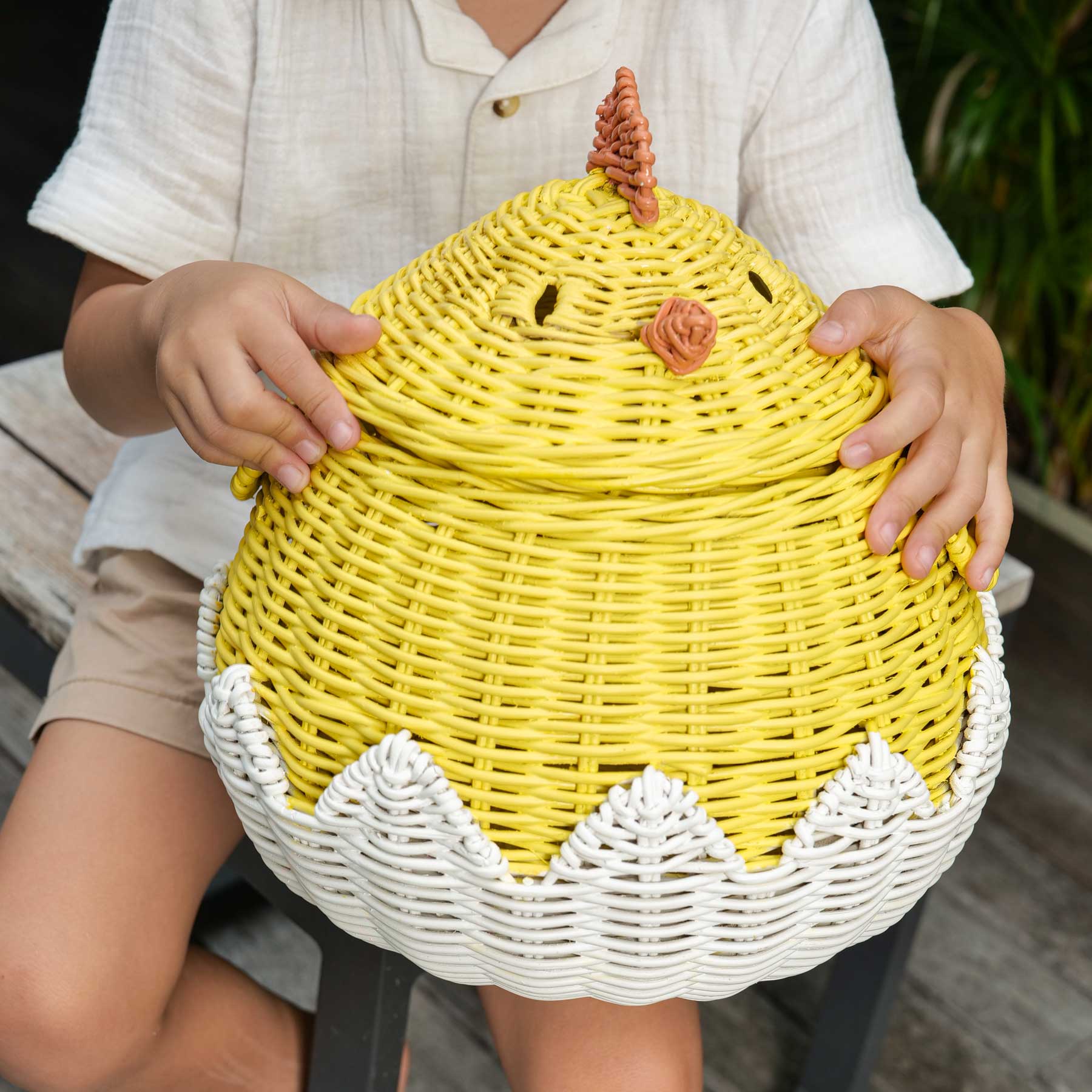 Child holding the handcrafted yellow rattan wicker chick storage basket by MOMIJI, featuring the orange beak and comb, and hite cracked eggshell base.