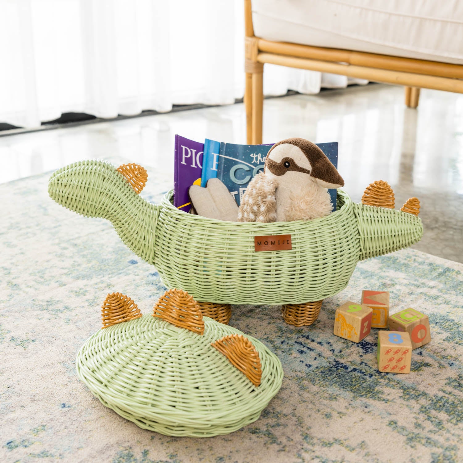 A medium size handcrafted dinosaur storage basket by MOMIJI in mint green rattan, opened to show toys and children’s books inside with lid set beside, placed on a carpeted floor.