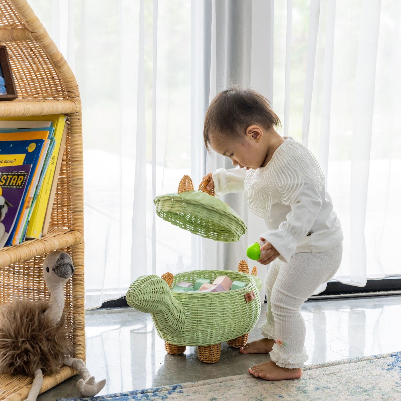 Child opening the lid of a medium size handcrafted dinosaur storage basket by MOMIJI in mint green rattan filled with colorful blocks, styled in a room with wicker shelves and soft curtains.