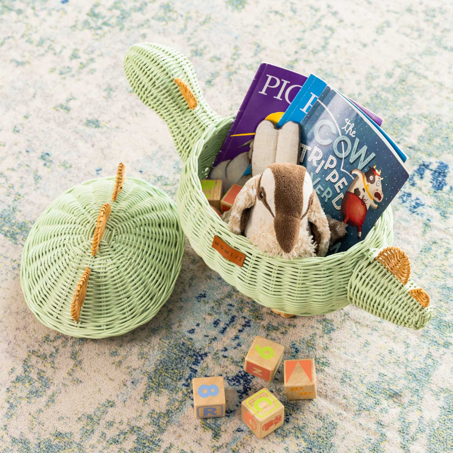 Top view of a medium size handcrafted dinosaur storage basket by MOMIJI in mint green rattan, opened to reveal toys and books with the lid set beside it on a carpet.