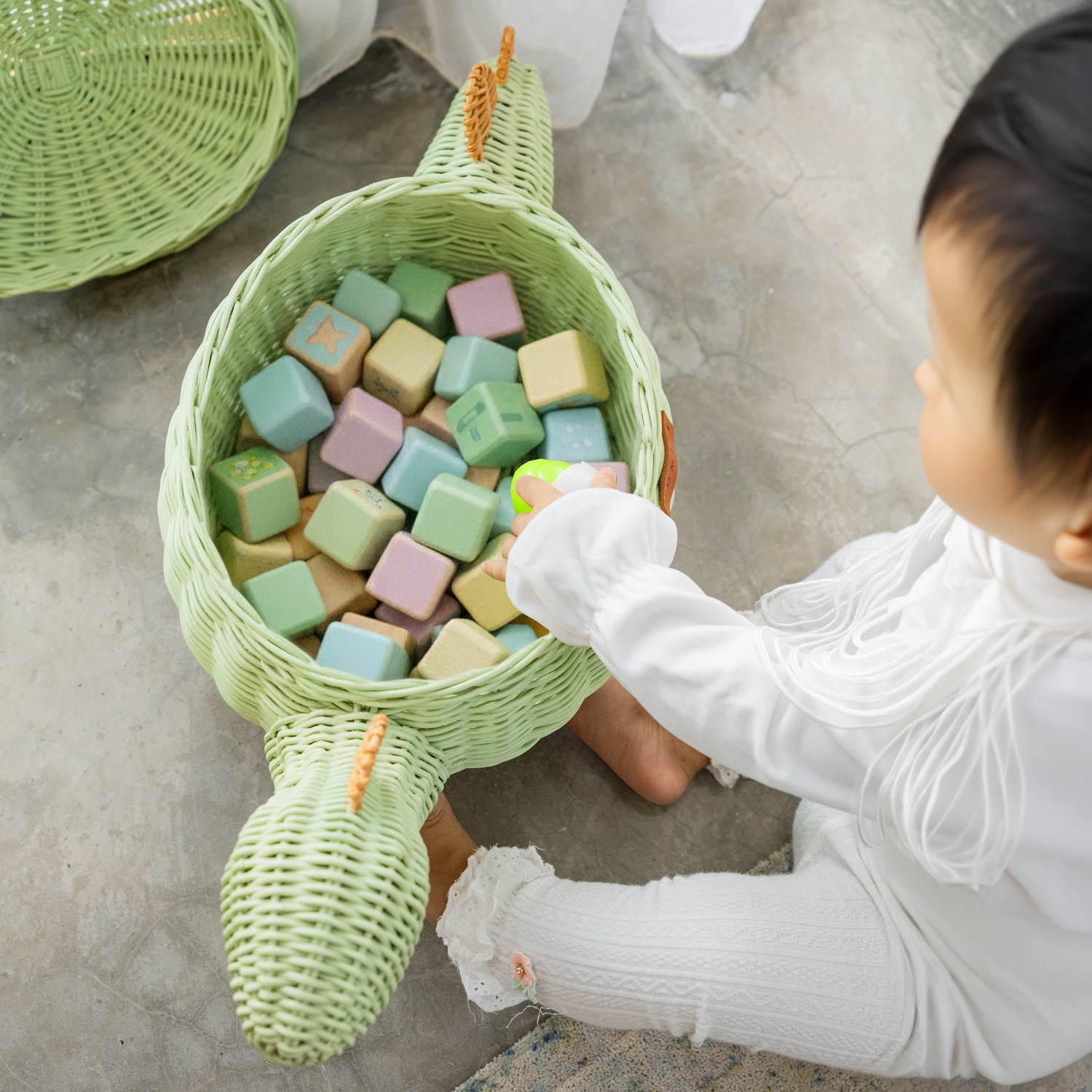Top view of a child reaching for colorful blocks inside a medium size handcrafted dinosaur storage basket by MOMIJI in mint green rattan, with the lid placed beside it on a concrete floor.