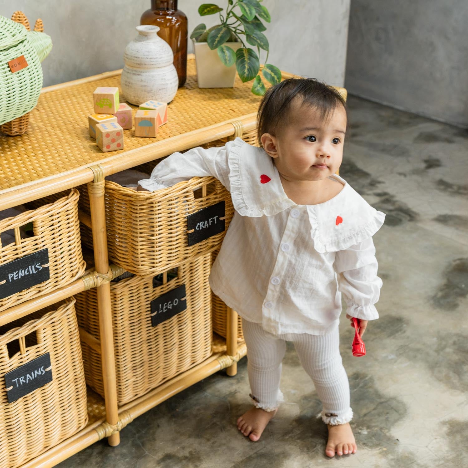 Child in a white outfit opening a basket on the Graha Montessori rattan storage shelf by MOMIJI, with blackboard-labeled baskets and styled decor on top.