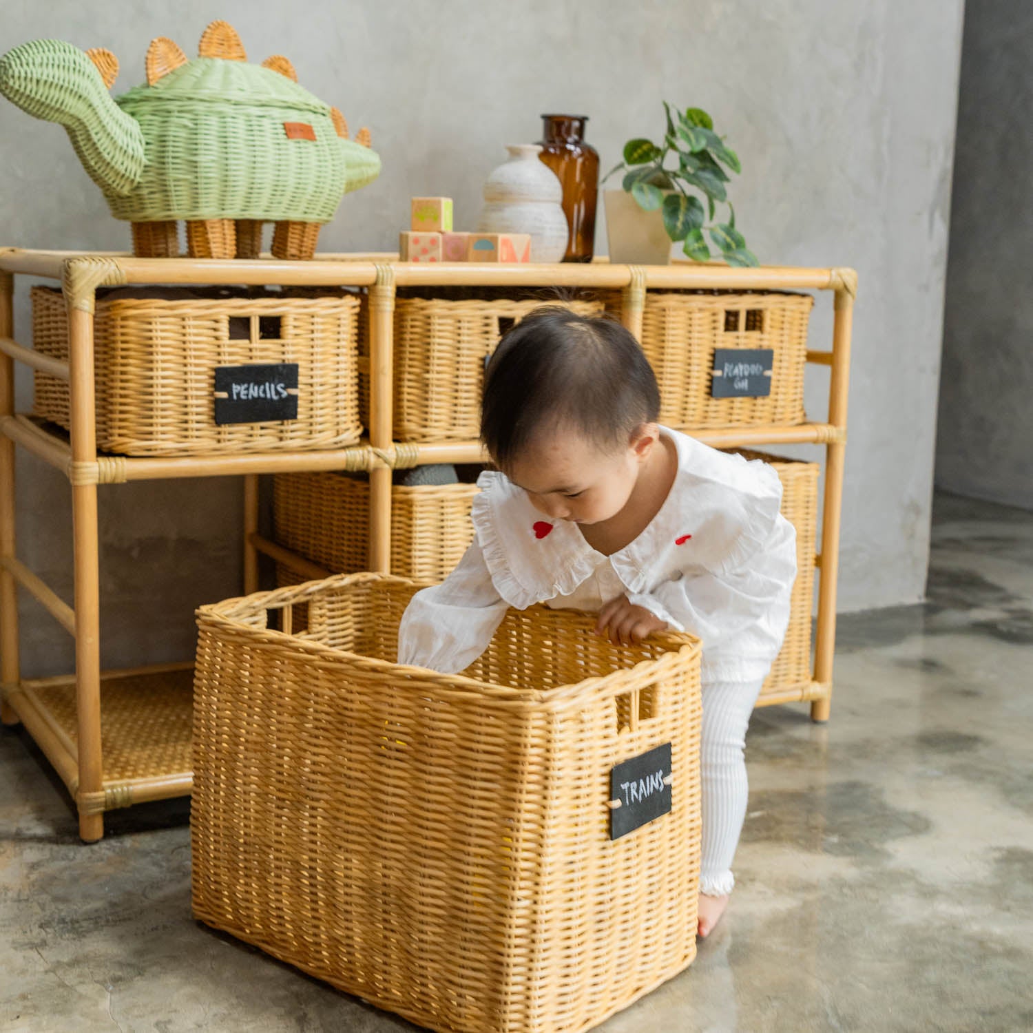 Child searching for toys in a wicker basket with a blackboard-labeled “Trains” tag from the Graha Montessori shelf by MOMIJI, in a room with a mint green rattan dinosaur basket.
