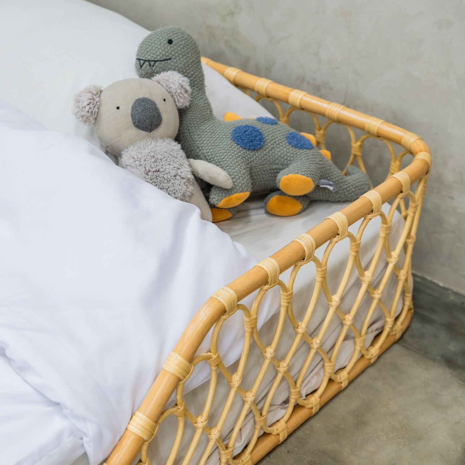 Two plush toys, a dinosaur and a koala, placed in the corner of the handcrafted Layla Montessori rattan floor bed by MOMIJI in natural beige.