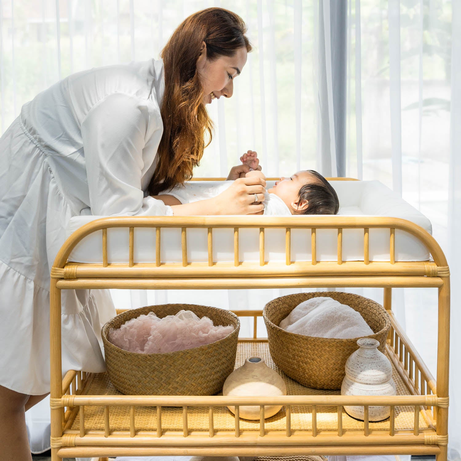 Woman interacting with a baby laying on the handcrafted rattan wicker Lestari changing table with shelves by MOMIJI, with baby's item on the shelf.