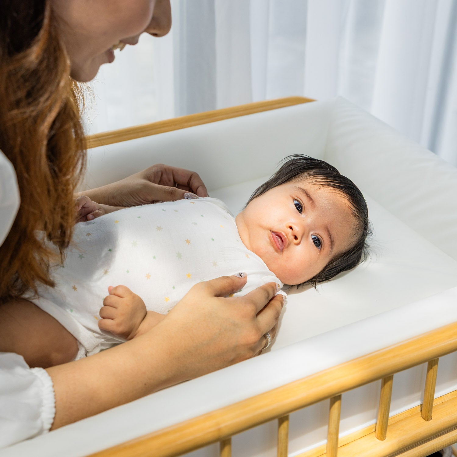 Baby lying on the handcrafted rattan wicker Lestari changing table with shelves by MOMIJI with woman beside in a calm indoor setting.