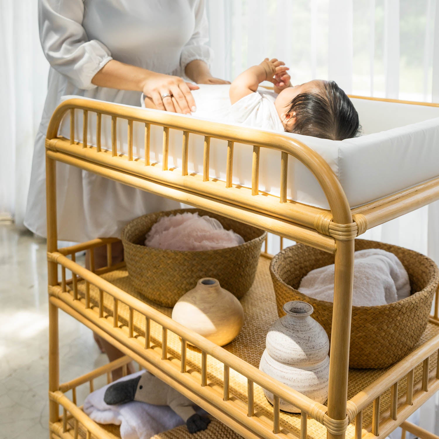 Baby lying on the handcrafted Lestari rattan changing table and shelves by MOMIJI, with angled view highlighting the rattan frame and rattan wicker shelf bases.