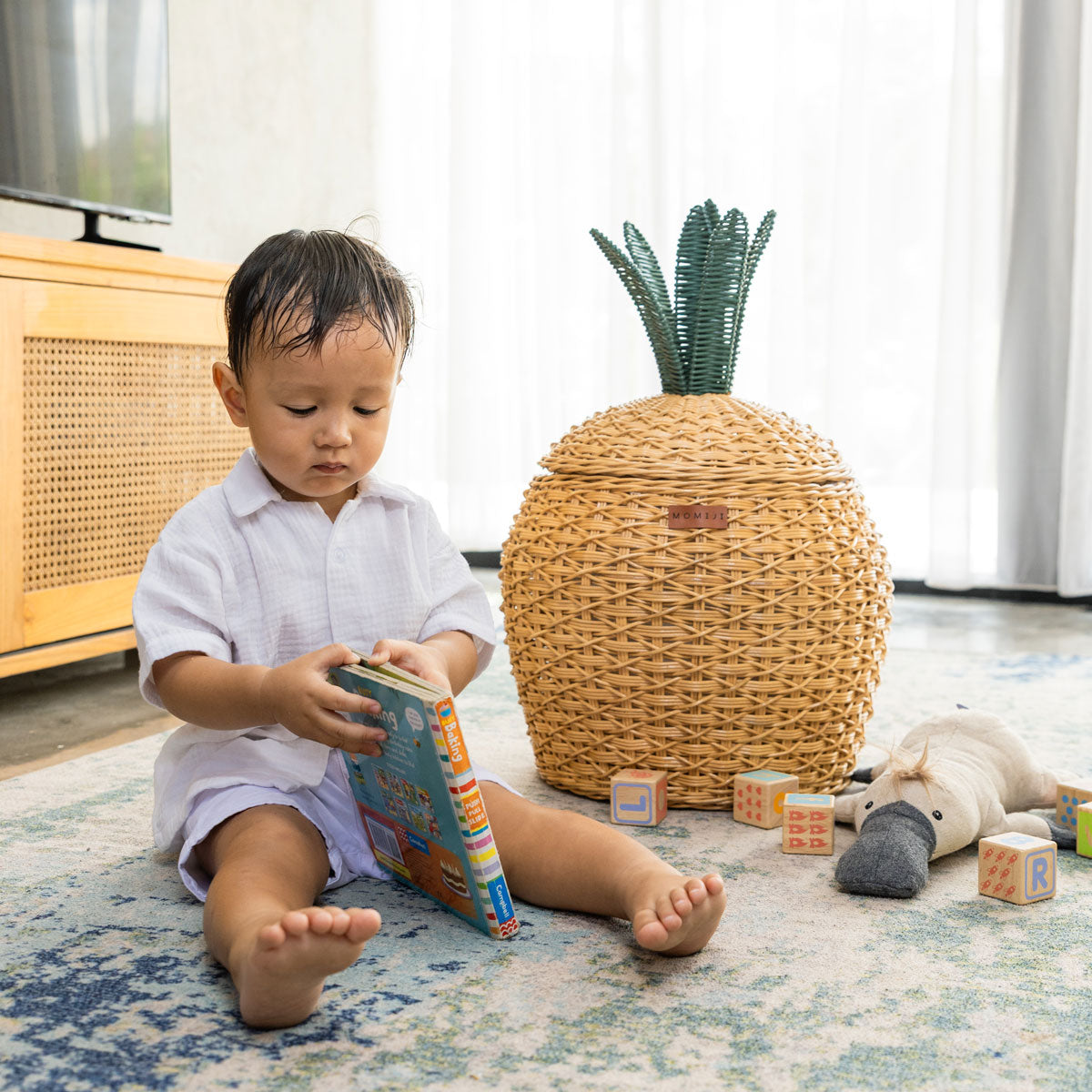 Child sitting on a rug reading a book beside a handcrafted natural rattan wicker pineapple storage basket in medium size by MOMIJI, styled with toys in a calm play space.