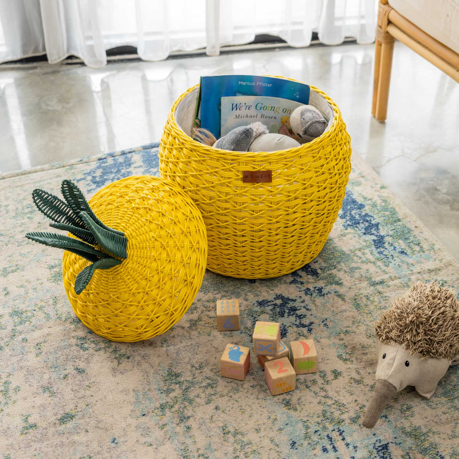 Handcrafted yellow rattan wicker pineapple storage basket in medium size by MOMIJI, shown with the lid off and filled with toys and children’s books on a a patterned rug, encouraging Montessori-inspired organization.