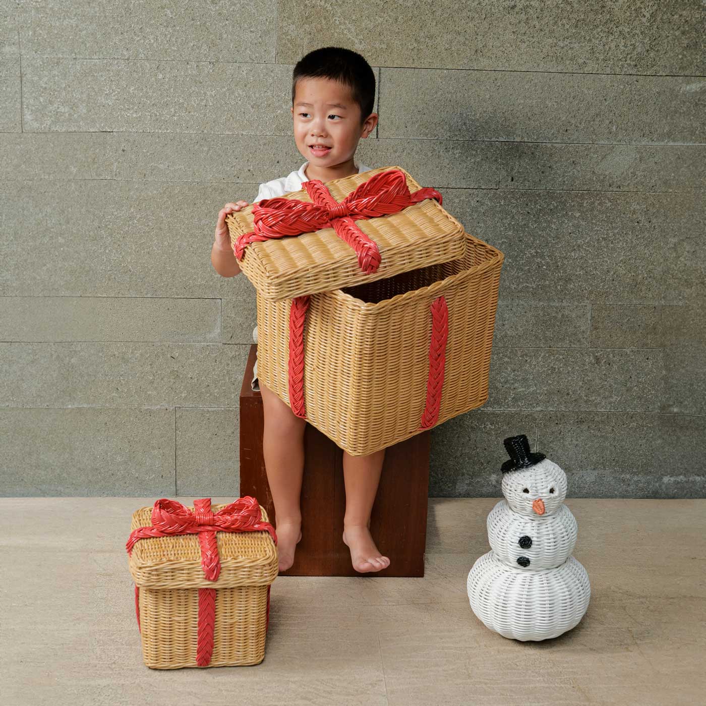 Child holding a large sized of the handcrafted rattan wicker storage gift box by MOMIJI with snowman rattan basket and small sized rattan basket gift box on the floor.