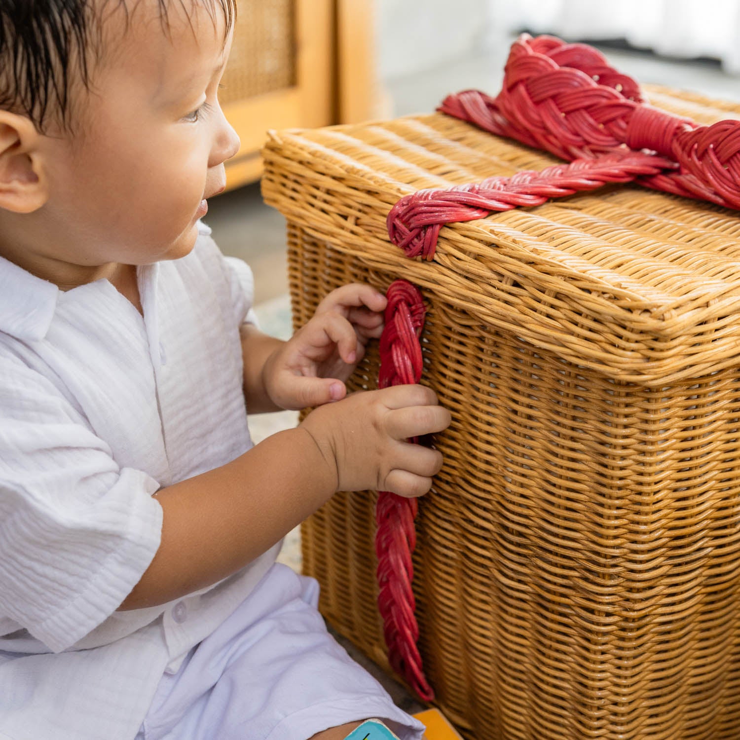 Child holding the red ribbon line on the side of a handcrafted rattan wicker gift box by MOMIJI, showing the decorative wrap-around detail.