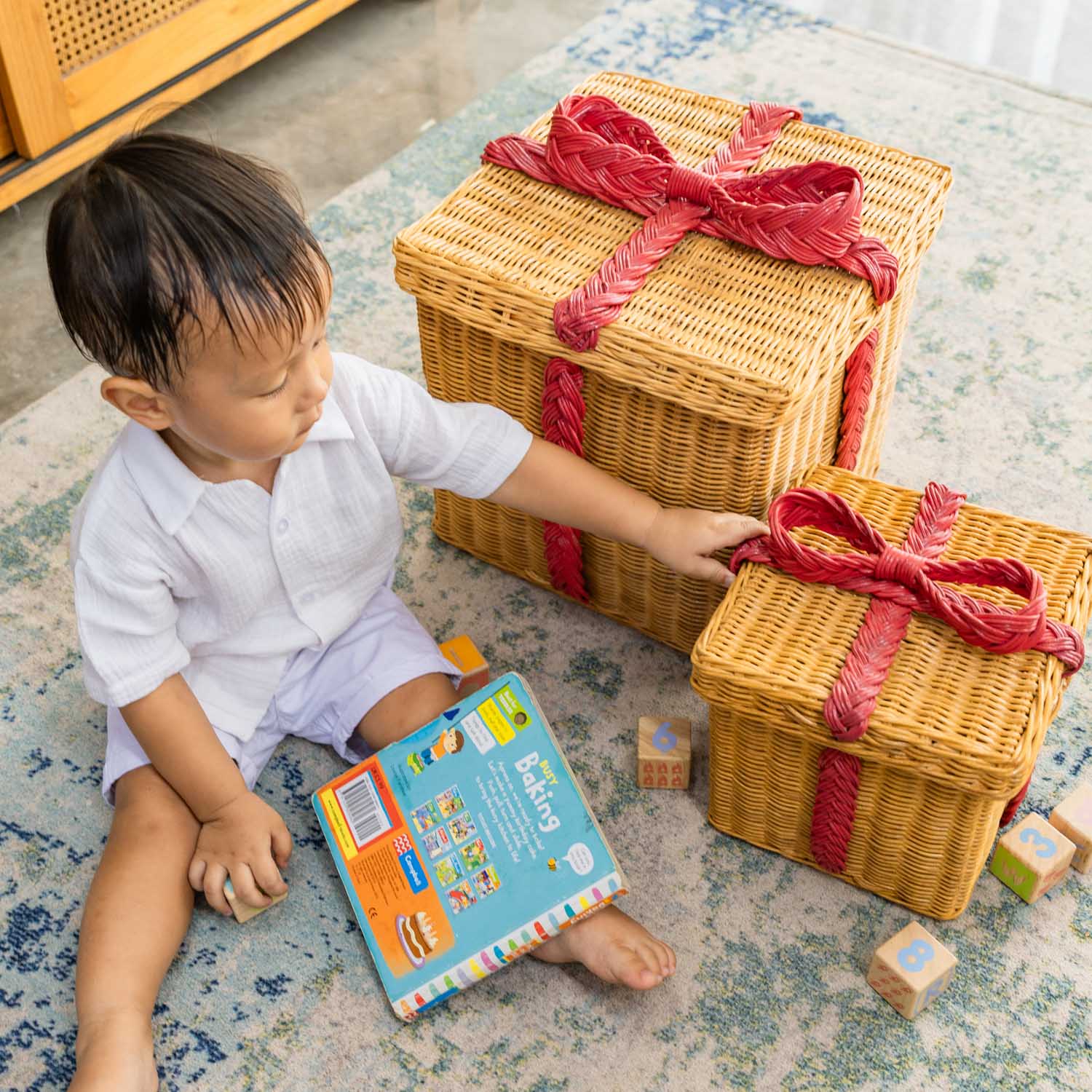 Child sitting on a patterned rug playing with handcrafted rattan wicker storage gift boxes by MOMIJI, holding the smaller box beside toys and books.