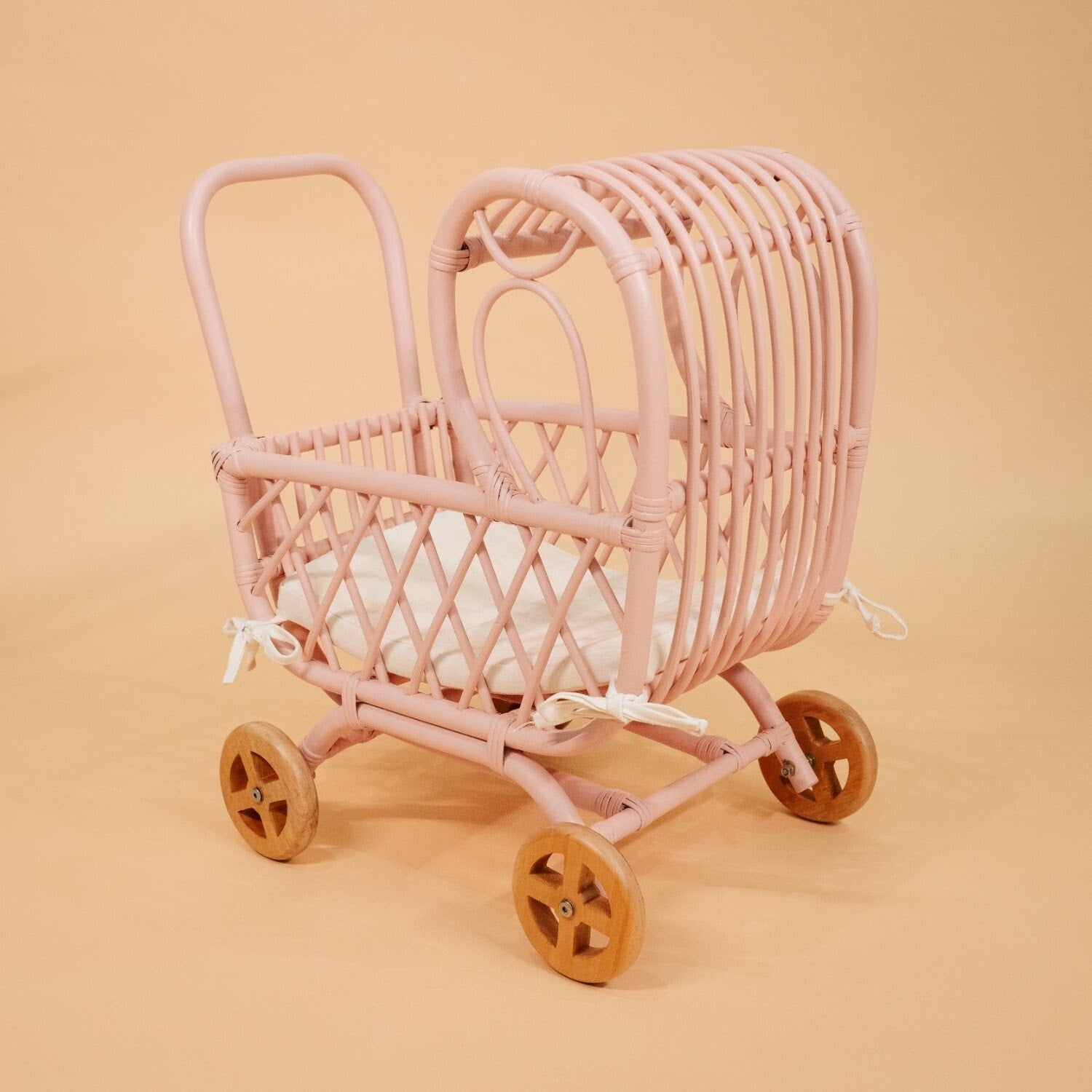 Side view of the handwoven rattan Layla Toy Stroller by MOMIJI in pink, with a white seat pad inside.