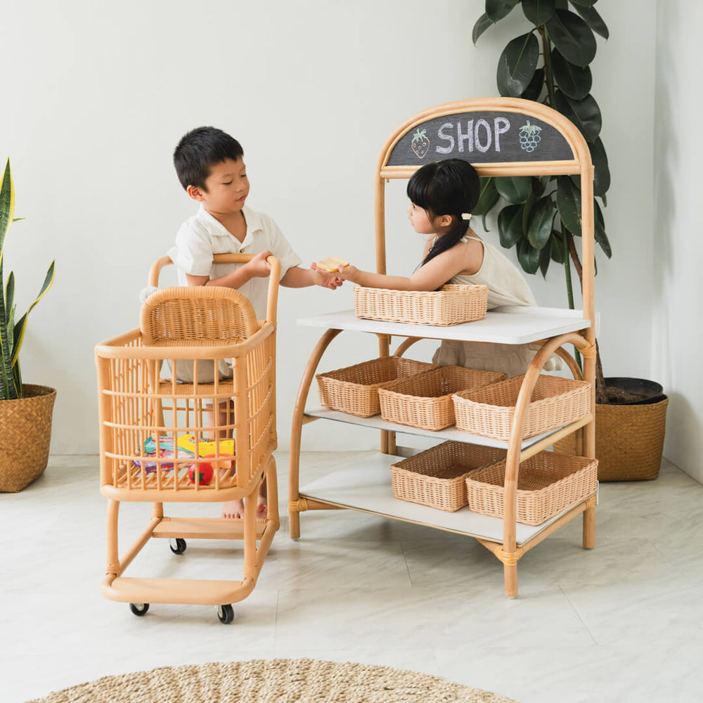 Two children playing shop using the 3-in-1 furniture by MOMIJI with an extra rattan shopping trolley.