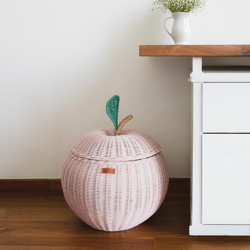 A pink apple rattan storage basket by MOMIJI next to a wooden table cabinet with a vase with flowers on top.