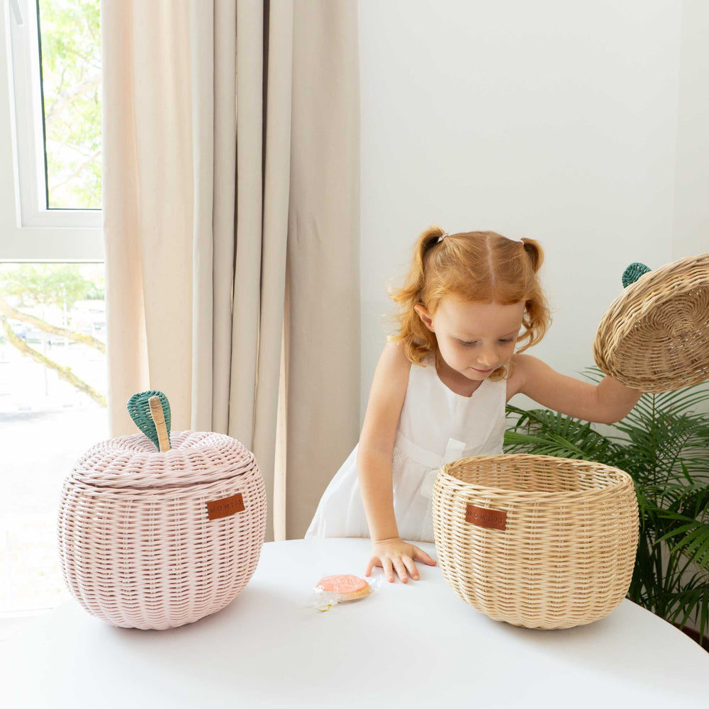Child opening the natural apple rattan storage basket while holding the lid checking what's inside the basket next to a pink apple rattan storage basket by MOMIJI.