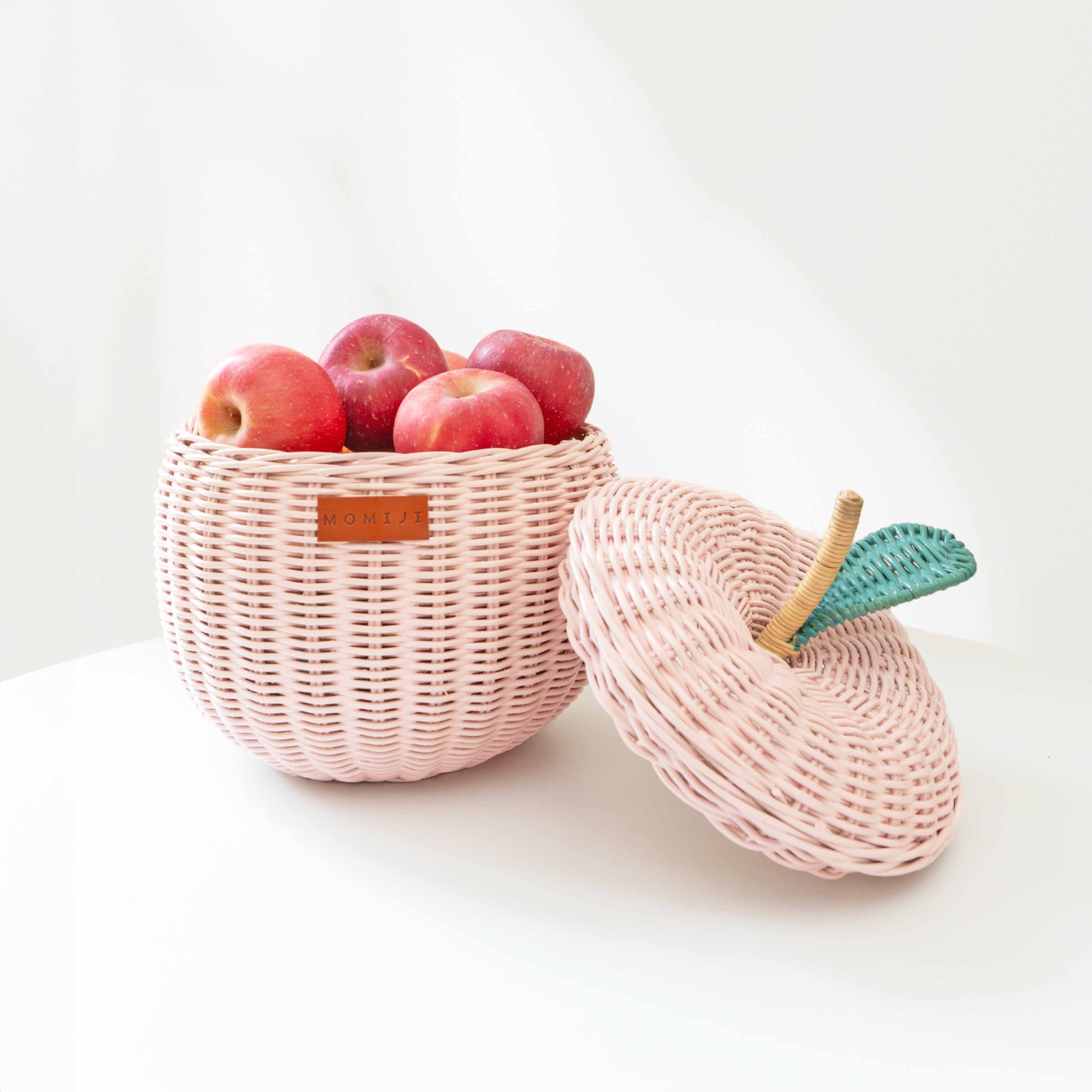 An opened small sized pink apple rattan storage basket by MOMIJI, full with fresh apples inside the basket with lid leaning beside the rattan basket.