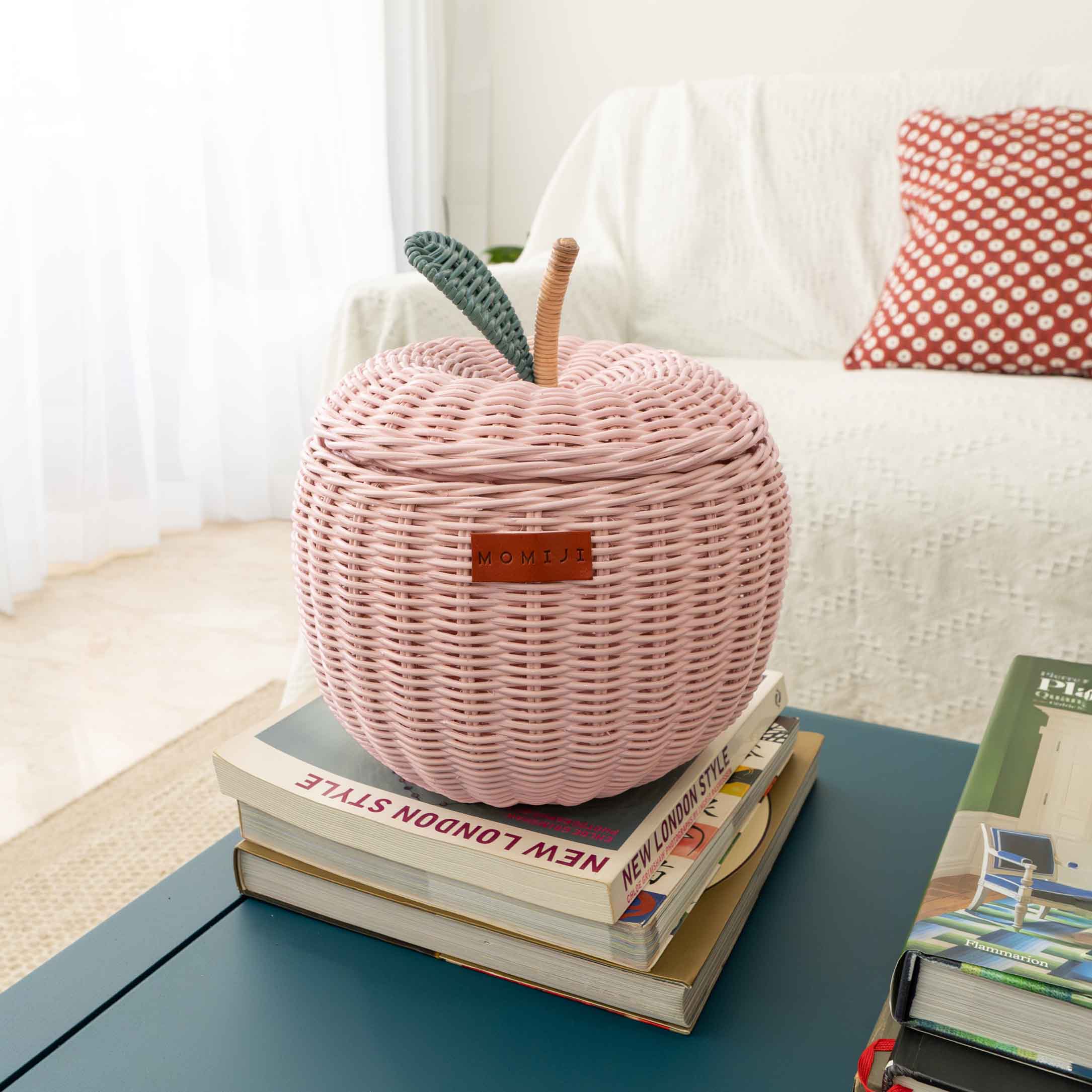 Pink woven apple rattan storage basket by MOMIJI on a stack of books with a white sofa and red pillow in the background.