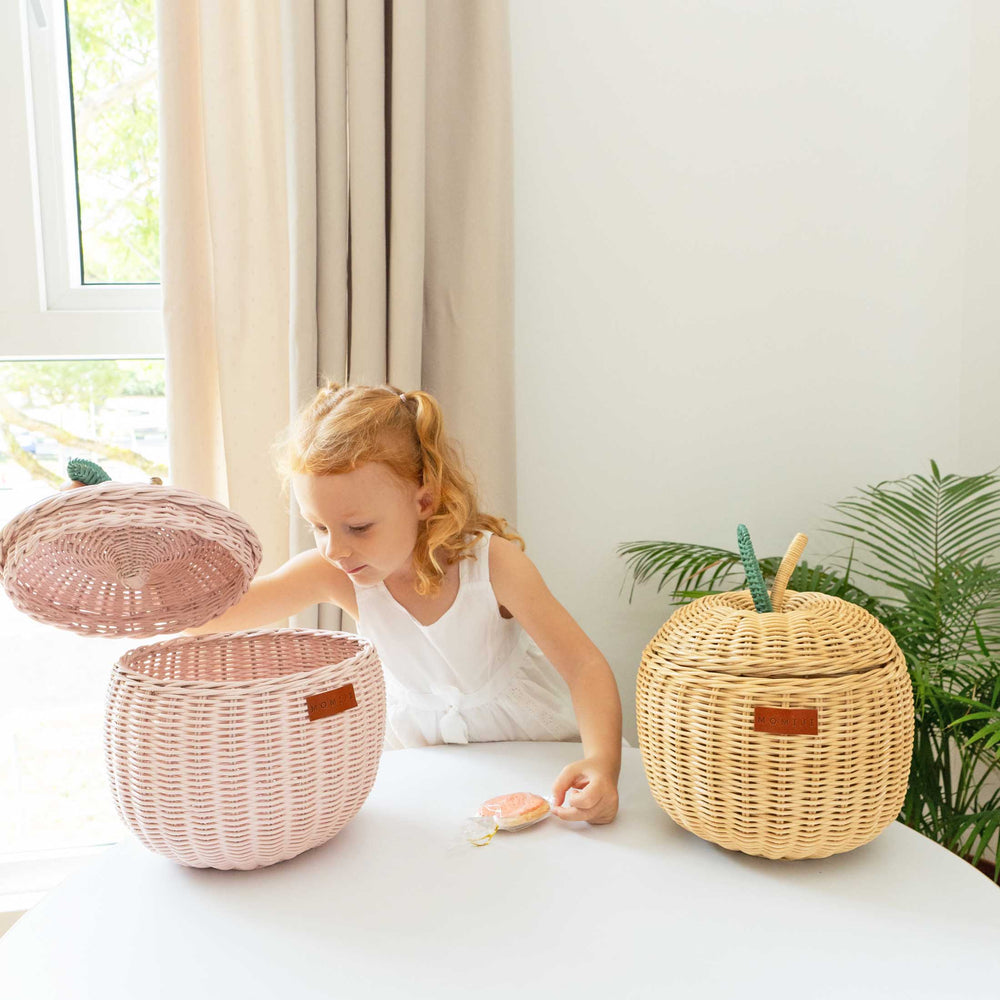 Child playing with the pink apple rattan storage baskets next to a natural apple rattan storage basket by MOMIJI in a bright room with plants.