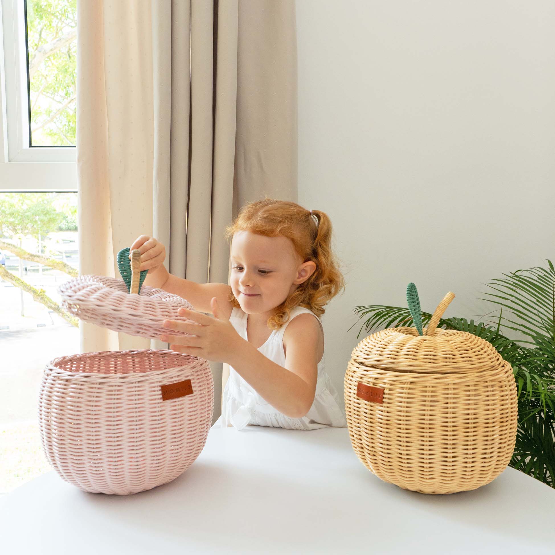 Child opening the pink apple rattan storage basket with two hands next to a natural apple rattan storage basket by MOMIJI.