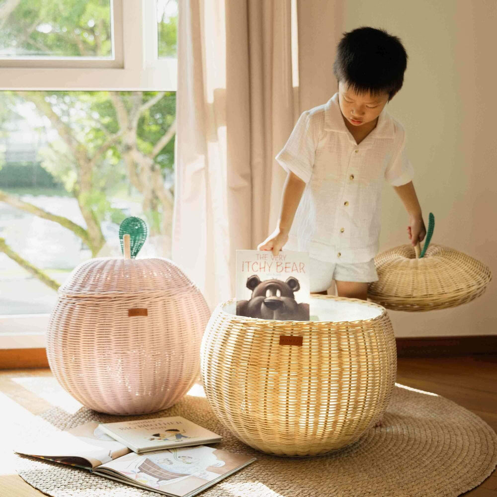 Child opening the natural apple rattan storage basket while holding the lid and picking up a book inside the rattan basket beside the pink apple rattan storage basket by MOMIJI.