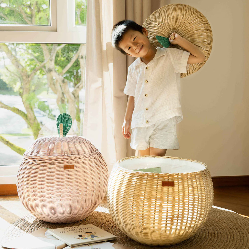 Child opening the natural apple rattan storage basket while holding the lid beside the pink apple rattan storage basket by MOMIJI.