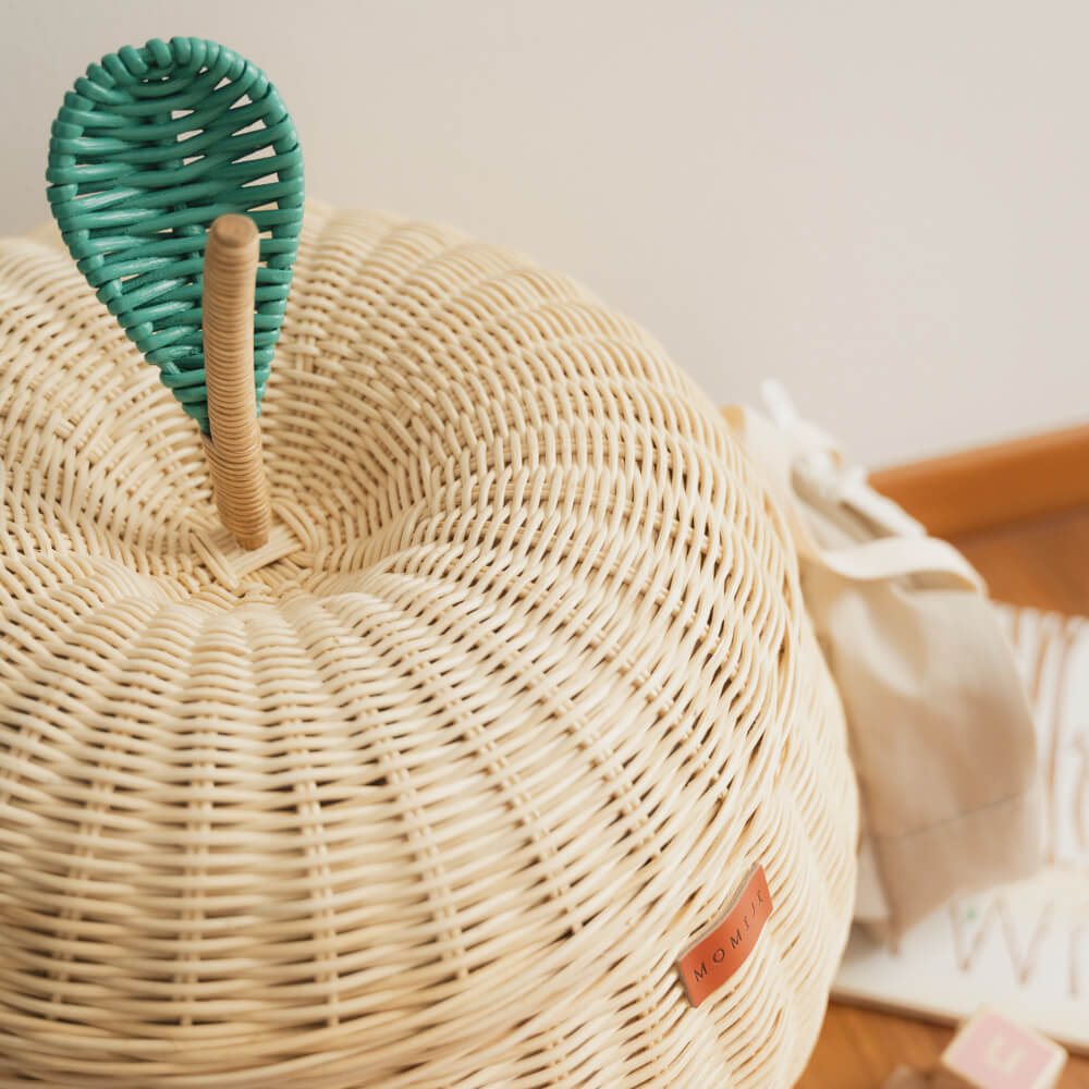 Close up view of natural apple rattan storage basket by MOMIJI with a green lid that looks like an apple leaf, on a wooden floor.