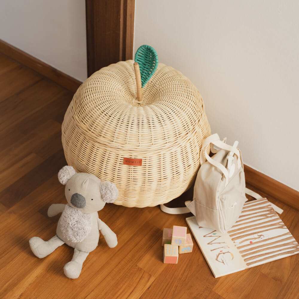 Top view of a natural apple rattan storage basket by MOMIJI with a green lid that looks like an apple leaf, on a wooden floor next to a stuffed toy, mini bag, a book, and wooden blocks. 