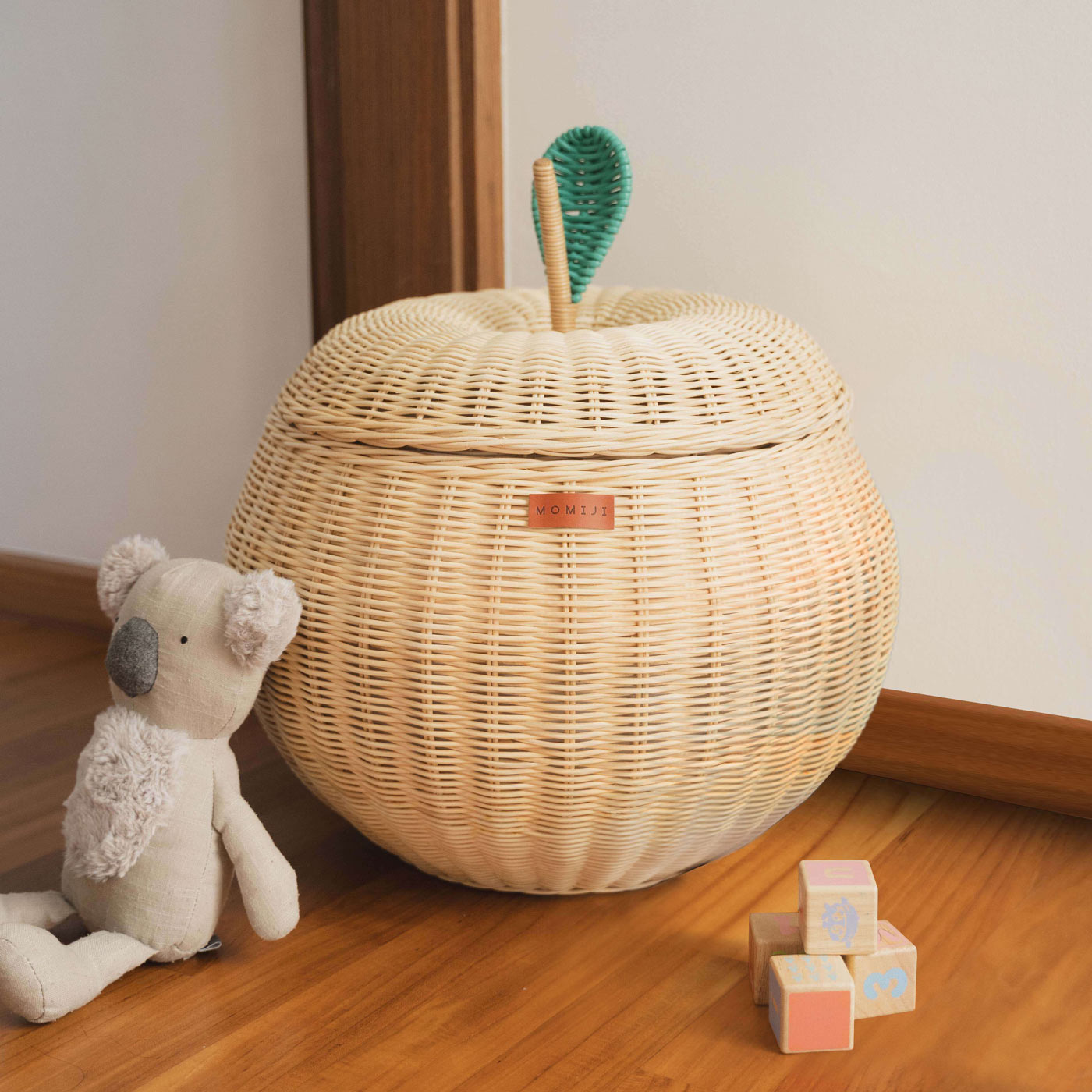 A natural apple rattan storage basket by MOMIJI with a green lid that looks like an apple leaf, on a wooden floor next to a stuffed toy and wooden blocks.