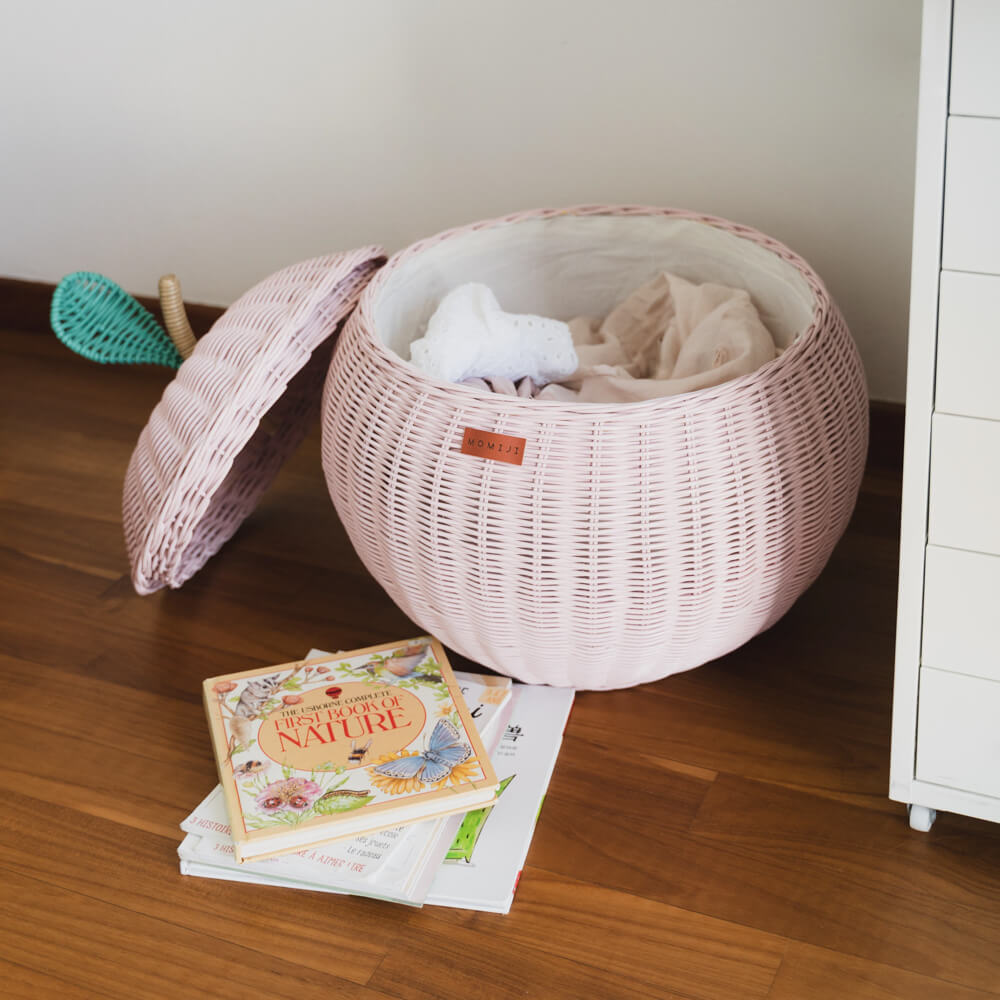 An opened pink apple rattan storage basket by MOMIJI with lid leaning beside the basket, with unfolded clothes inside the rattan basket on a wooden floor next to books.