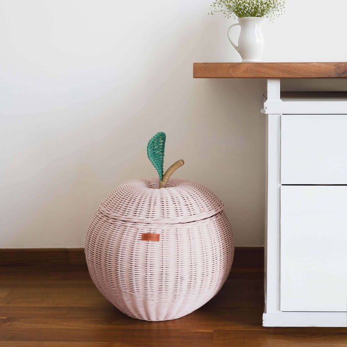 A pink apple rattan storage basket by MOMIJI next to a wooden table cabinet with a vase with flowers on top.