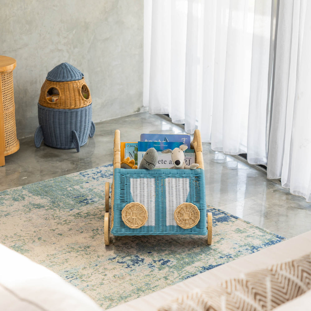 Wicker Archie Storage Wagon by MOMIJI filled with books and toys on a rug in a room with rattan furniture.