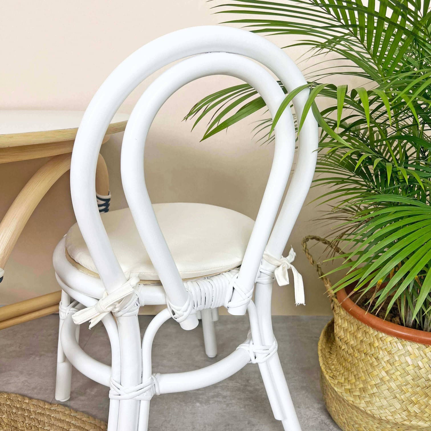 Back view of the Aulia Kids Chair by MOMIJI in an indoor setting with a rattan table and a potted plant, highlighting the white rattan frame.