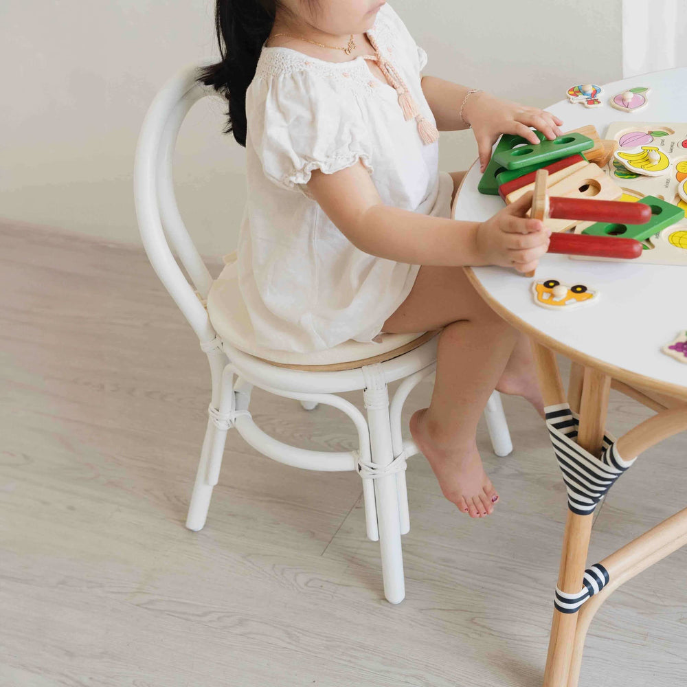 Child sitting on the Aulia Kids Chair by MOMIJI while playing with toys on a rattan table indoors, showcasing the white rattan frame.