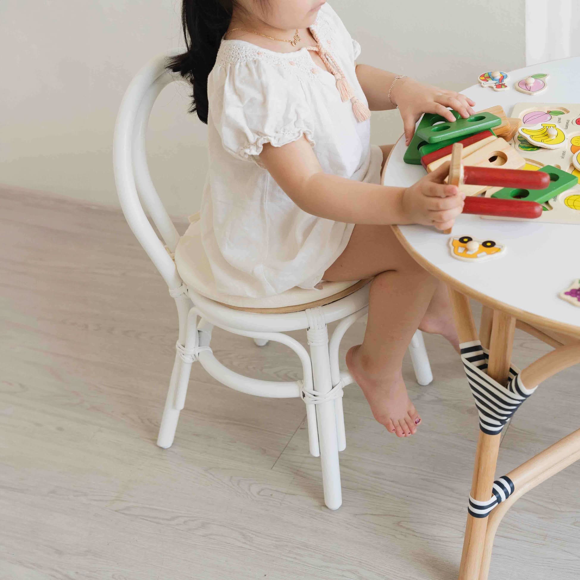 Child sitting on the Aulia Kids Chair by MOMIJI while playing with toys on a rattan table indoors, showcasing the white rattan frame.