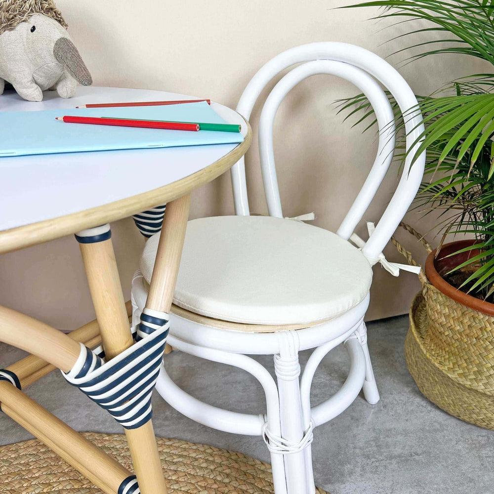 Aulia Kids Chair by MOMIJI with a white cushion placed beside a rattan table styled with books and toys in a room with plants.