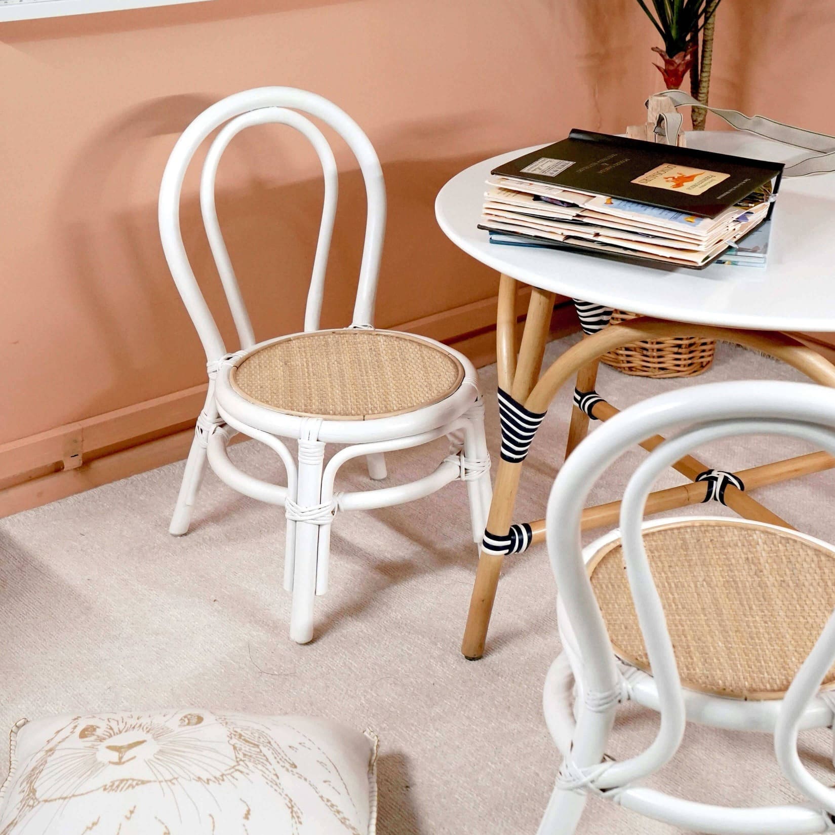 Two Aulia Kids Chairs by MOMIJI without cushions, featuring white rattan frames and natural handwoven rattan seats, placed on a rug beside arattan table with books in an indoor setting.