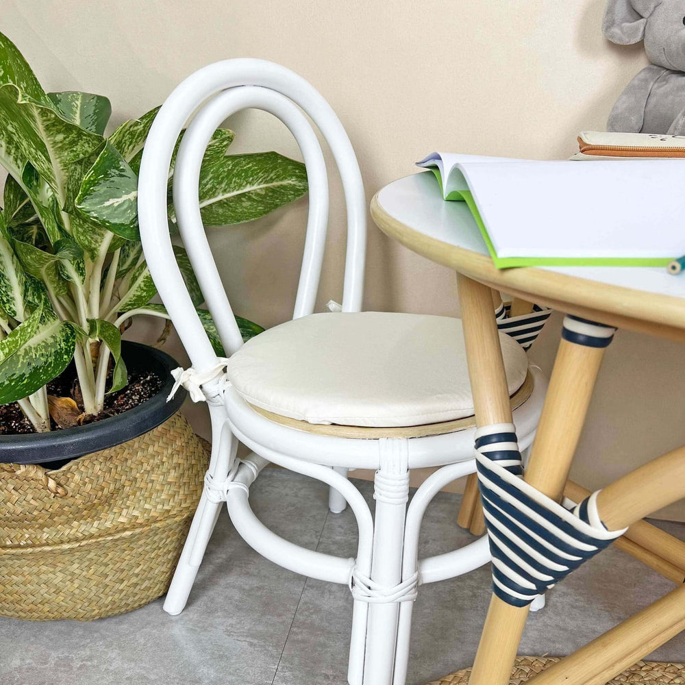 Aulia Kids Chair by MOMIJI with a white cushion placed in front of a rattan table in a room with plants, highlighting the white rattan frame.