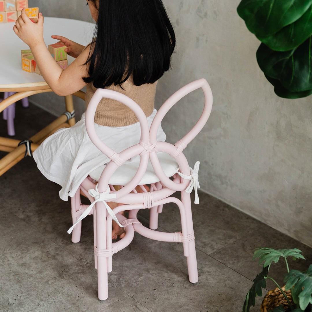 Back view of a child sitting on a handcrafted pink rattan wicker Butterfly Kids Chair by MOMIJI, playing colorful blocks at an Aulia Kids Table in a playful children's room.
