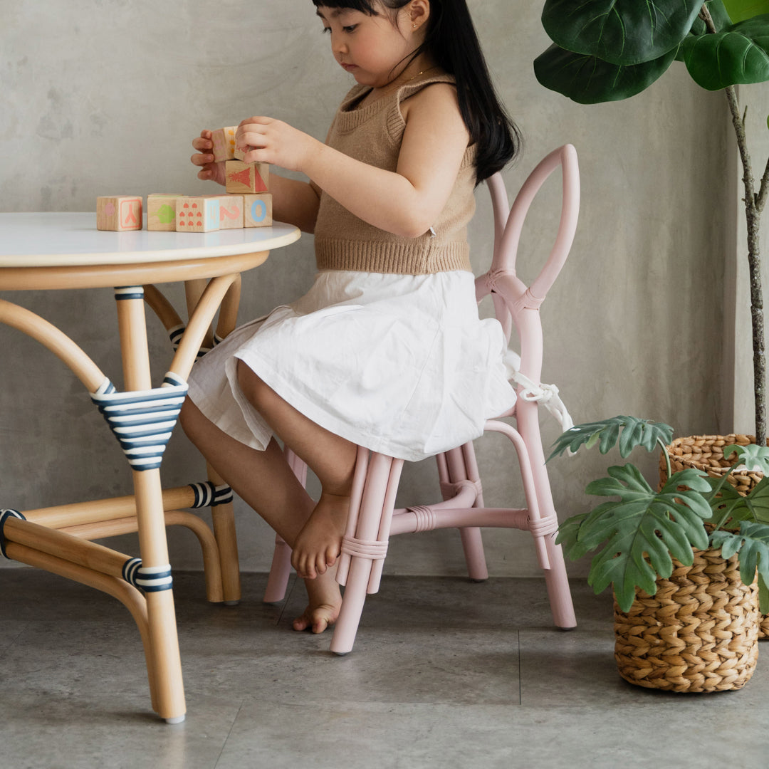 Child sitting on a handcrafted pink rattan and wicker Butterfly Kids Chair by MOMIJI, playing with toys at an Aulia Kids Table in a playful indoor setting.