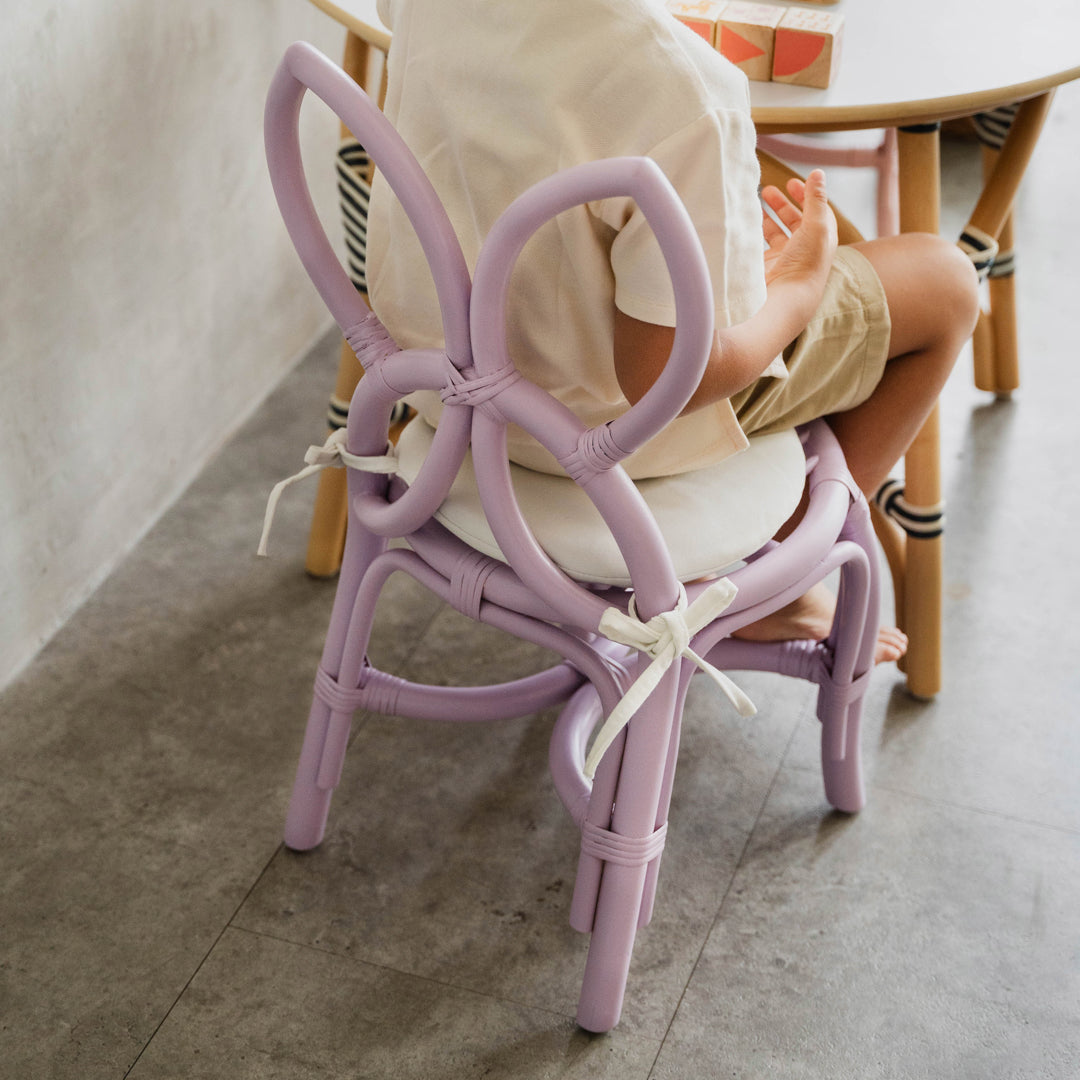 Back view of a child sitting on a handcrafted purple rattan wicker Butterfly Kids Chair by MOMIJI, playing at an Aulia Kids Table in a playful children's room.