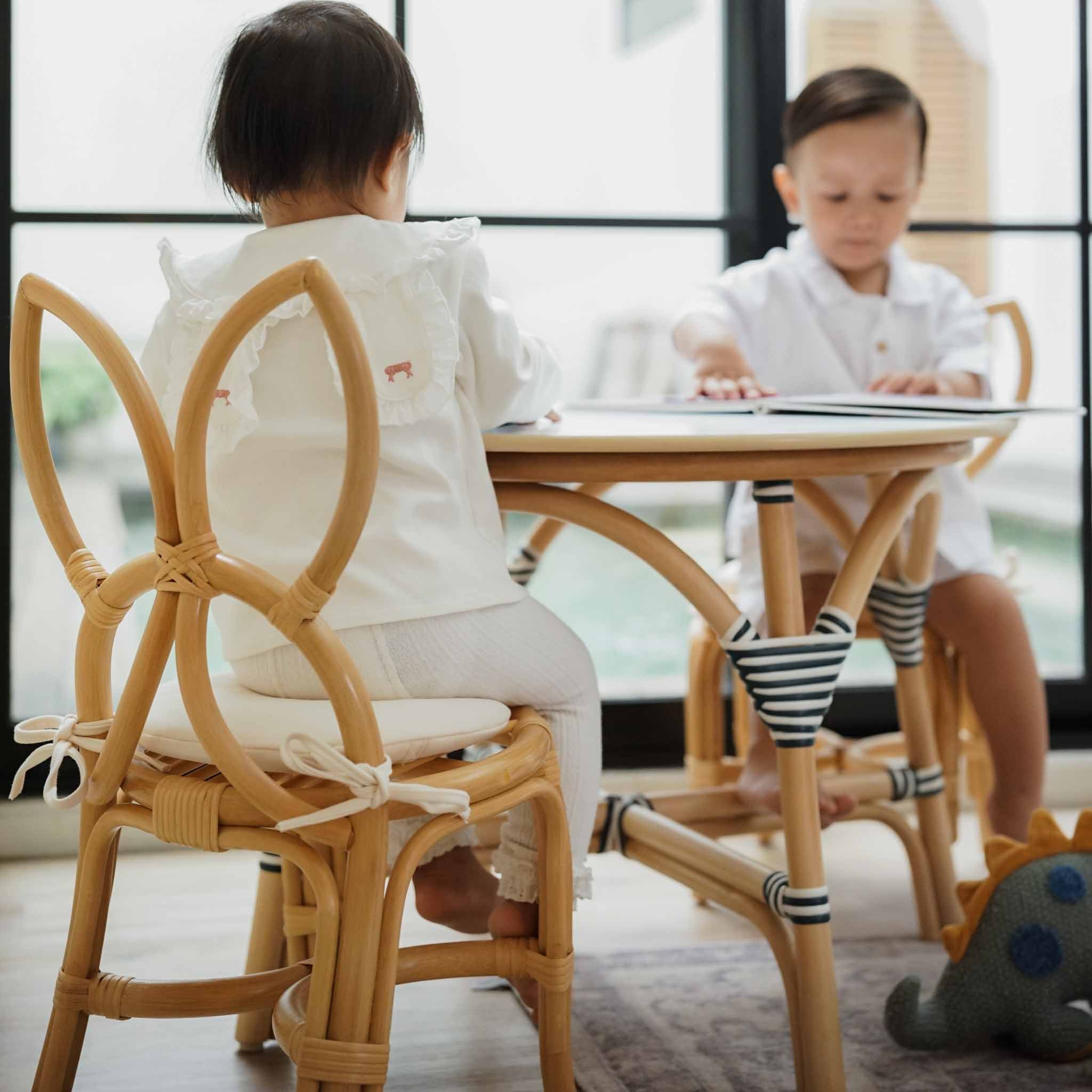 Two children playing and reading together using a handcrafted rattan wicker Aulia Kids Table (Medium) & Set of 2 Butterfly Kids Chairs (Natural) by MOMIJI for playroom decor.