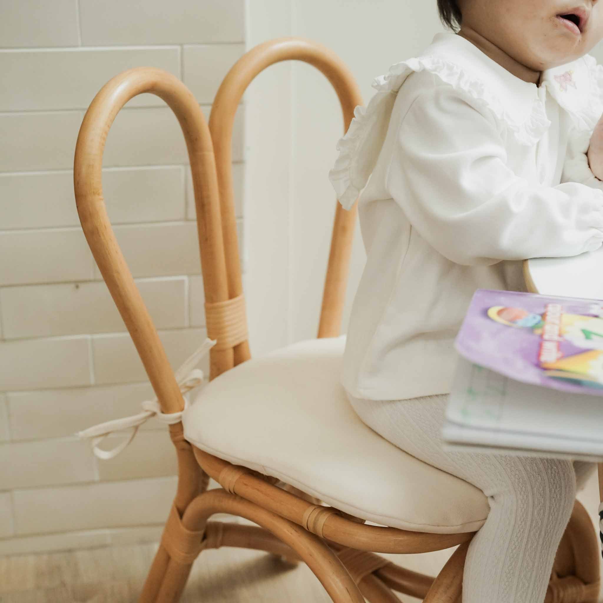 A child sit in the handcrafted rattan wicker Bunny Kids Chairs (Natural) by MOMIJI feature signature bunny-ear backrests with comfortable cushion seats. 