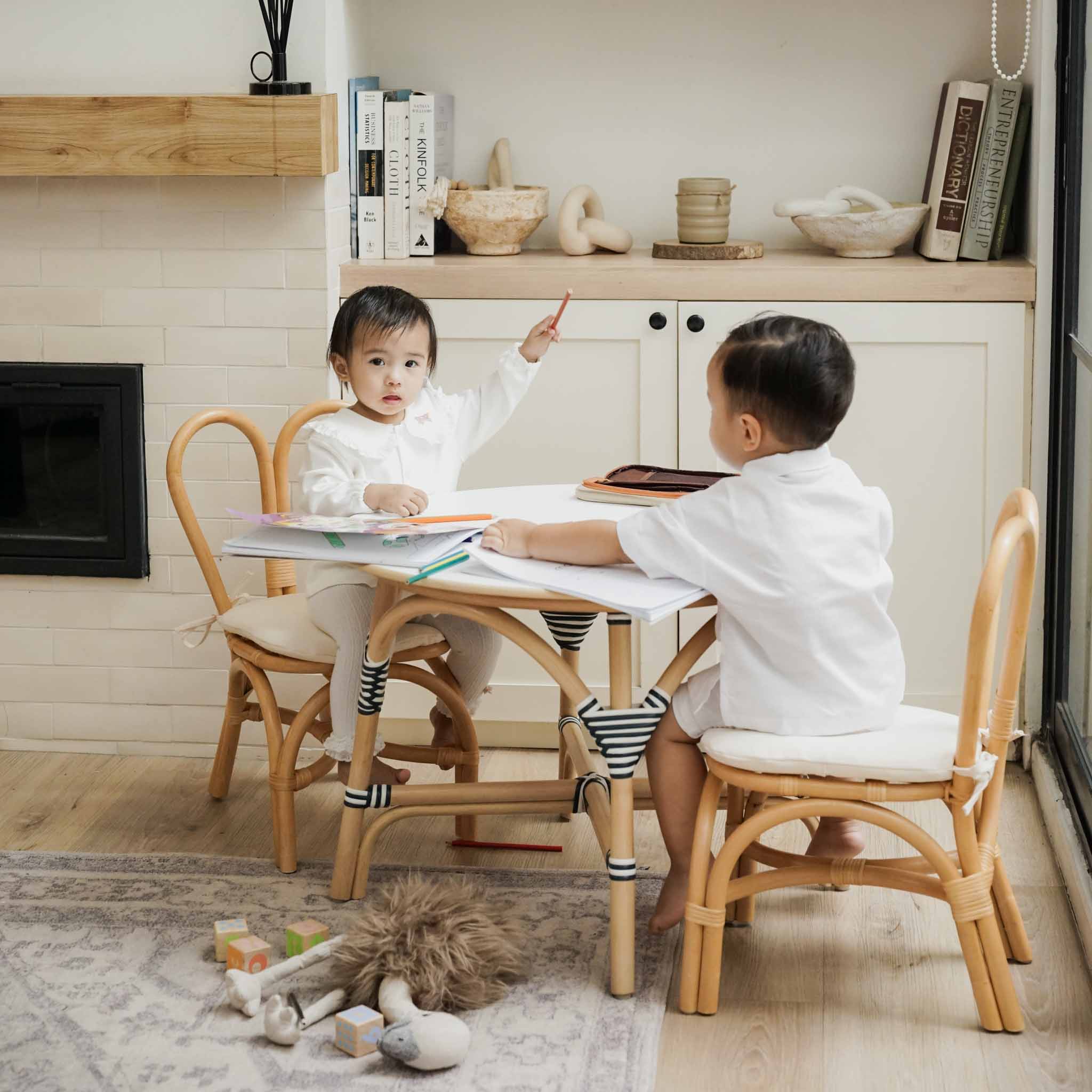 Two children sit at the Aulia Kids Table (Medium), paired with a Set of 2 Kids Bunny Chairs (Natural) by MOMIJI featuring handcrafted natural rattan frames, bunny-ear backrests, and soft cushioned seats. The Montessori-inspired children's furniture set is styled as a bright learning and activity space.